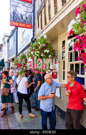 Due alti uomini in piedi in chat e azienda bicchieri da birra con altre persone che stanno dietro al di fuori città inglese public house, con fiori appesi in basso. Foto Stock