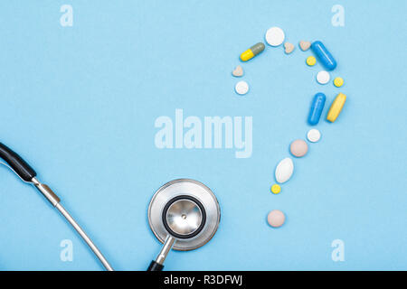 Varie pillole e capsule a forma di punto interrogativo e di segno phonendoscope su sfondo blu. Vista superiore Foto Stock