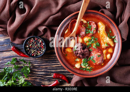 Fabada Asturiana fagiolo stufato caloroso in un vaso di argilla con il cucchiaio su una tavola in legno rustico con panno marrone, cucina spagnola, vista da sopra, flatlay, clo Foto Stock