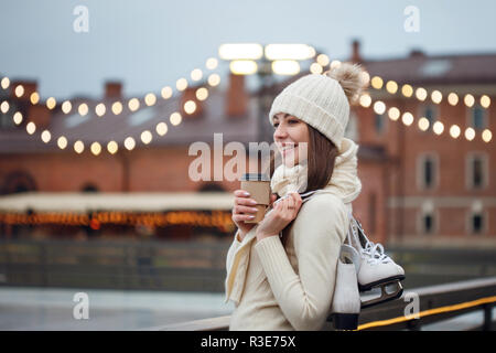 Felice giovane donna in maglia maglione e hat sta andando il pattinaggio. Giovani brunette con pattini sulla sua spalla, City Park in inverno, la pista di pattinaggio su ghiaccio. Foto Stock