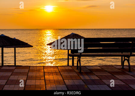 Vista in mattina presto Sun, dawn, sulla spiaggia pubblica con ombrelloni di paglia. Panca in legno è collocato sulla passerella piastrellato. Foto Stock