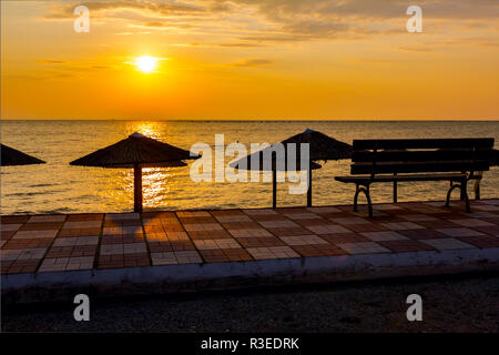 Vista in mattina presto Sun, dawn, sulla spiaggia pubblica con ombrelloni di paglia. Panca in legno è collocato sulla passerella piastrellato. Foto Stock