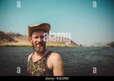 Candida fotografia dell'uomo degli anni trenta sorridendo e guardando gioioso alla macchina fotografica, in piedi accanto al fiume. L'aria è fumosa a seguito di incendi boschivi. Foto Stock