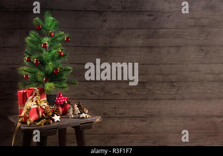 Albero di Natale artificiale con red baubles confezione regalo e decorazione su un piccolo tavolo di fronte un rustico di pareti in legno, generoso spazio di copia selezionato, Foto Stock