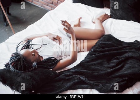 Bella ragazza africana in sleepwear sorridente ascoltando la musica nelle cuffie giacente sul letto Foto Stock