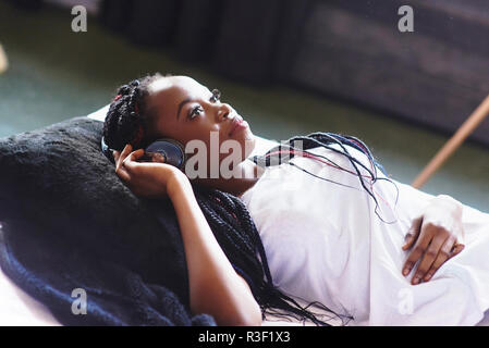 Bella ragazza africana in sleepwear sorridente ascoltando la musica nelle cuffie giacente sul letto. Gli occhi chiusi. Da sopra Foto Stock