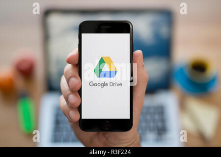 Un uomo guarda al suo iPhone che visualizza le unità di Google logo, mentre se ne sta seduto alla sua scrivania per computer (solo uso editoriale). Foto Stock