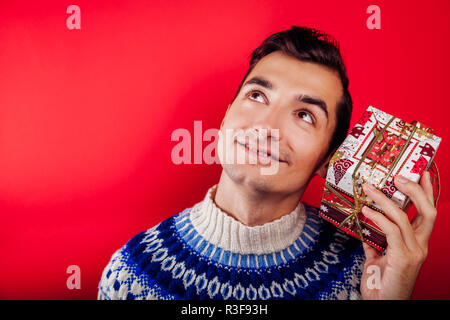 Studio shot di un giovane uomo in islandese maglione tenendo un regalo su sfondo rosso. Natale o Capodanno concetto. Foto Stock