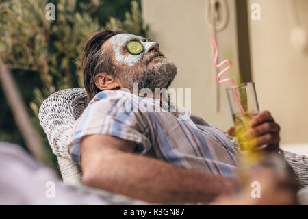 Rilassata anziani uomo seduto su una sedia con maschera per il viso sulla faccia tenendo un bicchiere di succo di frutta. Uomo in pensione rilassante con argilla maschera sul viso. Foto Stock