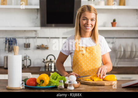 Felice giovane donna il taglio di vegetali e sorridente in telecamera in cucina Foto Stock