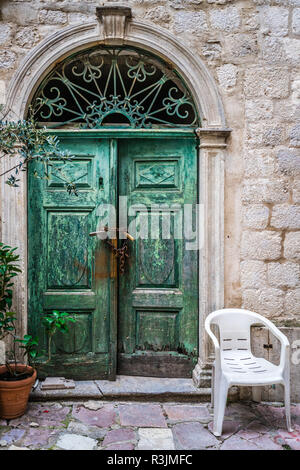 Sedia in plastica bianca di fronte bloccato con lucchetti e catene verde vecchio danneggiato porte in legno in uno degli edifici di Kotor, Montenegro Foto Stock