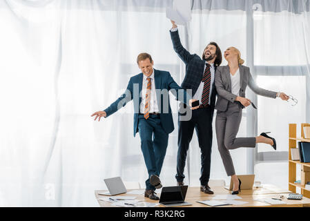 Assicurazione di gioiosa lavoratori ballando sul tavolo e gettando carte alla riunione in ufficio Foto Stock