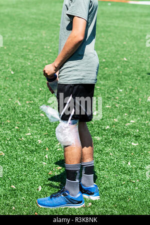 Una scuola di runner ha un avvolgimento di ghiaccio sul suo ginocchio mentre in piedi su un tappeto erboso verde campo pratica a guardare. Foto Stock