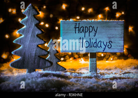 Cartello in legno con la calligrafia Inglese Buone Vacanze. Vintage albero di Natale sulla neve e le luci fairy in background. Spotlight blu Foto Stock