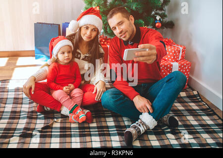 Buon Natale e Felice Anno Nuovo. Giovane uomo si siede sul letto accanto alla donna e bambino. Egli detiene il telefono e prende selfie. Donna e bambino lok e posa. Famiglia Foto Stock