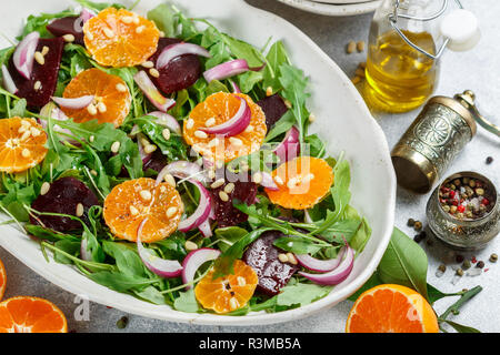Gourmet insalata di rucola, barbabietola cotta al forno, tangerini e cipolle rosse con spezie e pinoli in un piatto bianco. Olio di oliva e gli ingredienti su un livello di grigio sur Foto Stock