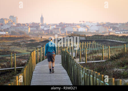 Uomo di mezza età passeggiate sul lungomare nel mezzo di vegetazione verso città lontana. Caucasici, Calvo, cappuccio nero, blu maglione grigio shorts. Vista posteriore. Copia Spac Foto Stock