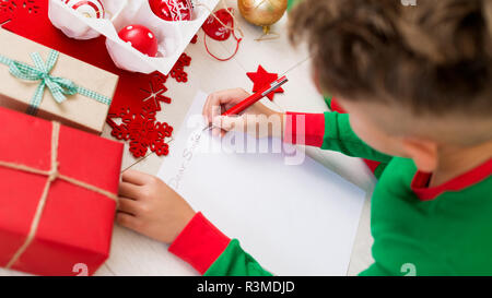 Carino il ragazzo che indossa il natale pigiami scrivere lettera a Babbo su livingroom piano. Vista aerea di un giovane ragazzo di scrivere il suo natale wishlist. Foto Stock