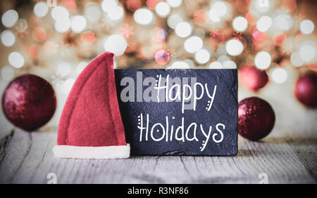 Piastra con la calligrafia Inglese Buone Vacanze. Vini spumanti e scintillanti luci dello sfondo. Decorazione di natale come sfere viola e Santa Hat Foto Stock