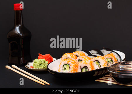 Uramaki California. Rotoli di Sushi con nori, riso, pezzi di avocado, cetriolo, decorata con pesci volanti roe su piastra ceramica. Bottiglia e ciotola con così Foto Stock