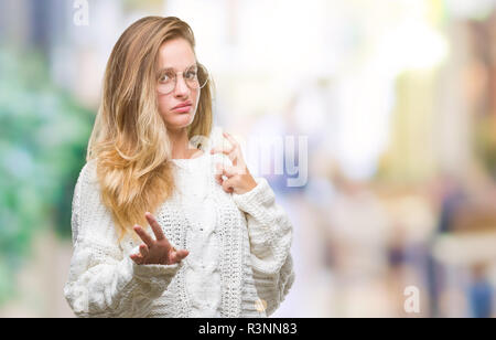 Giovane bella donna bionda che indossa un maglione invernale e occhiali da sole su sfondo isolato disgustato espressione, orinato e timorosi facendo disgusto Foto Stock