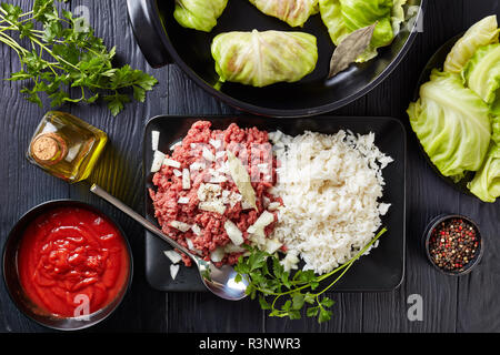 Materie ripieni di foglie di cavolo con ingredienti - le carni bovine macinate, riso bollito, il prezzemolo e la salsa di pomodoro su una tavola nera, vista da sopra, piana, laici, vicino-u Foto Stock