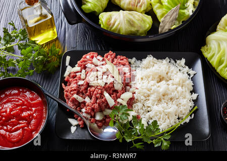 Materie ripieni di foglie di cavolo con ingredienti - le carni bovine macinate, riso bollito, il prezzemolo e la salsa di pomodoro su una tavola nera, vista da sopra,, close-up Foto Stock