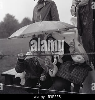 1965, storico, felice di spettatori sotto la pioggia, un allegro madre e figlia seduti insieme sotto un piccolo ombrello, riparo dai il tempo umido in corrispondenza di un outdoor evento equestre, Inghilterra, Regno Unito. Foto Stock