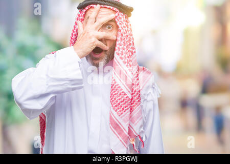 Senior uomo arabo indossando keffiyeh su sfondo isolato peeking in stato di shock che copre il viso e gli occhi con la mano, guardando attraverso le dita con imbarazzo Foto Stock