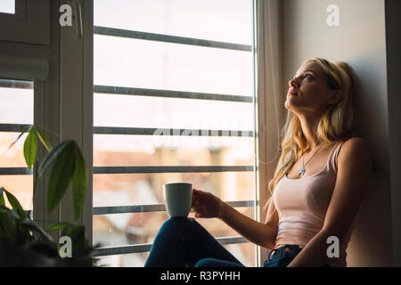 Giovane donna bionda holding tazza da caffè seduti nel davanzale Foto Stock