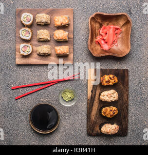 Varietà di rotoli su un piatto di legno con salsa di soia e zenzero, wasabi accanto giacciono le bacchette di colore rosso su sfondo rustico, vista dall'alto Foto Stock