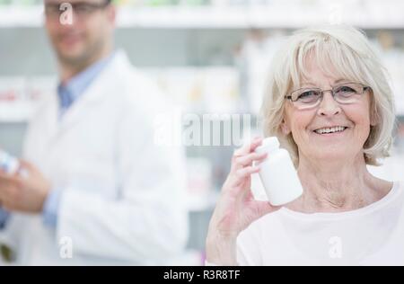 Ritratto di donna senior azienda pillola bottiglia in farmacia, con il farmacista in background. Foto Stock