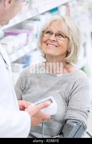 Il farmacista maschio controllo sorridente femmina senior di pressione del sangue del paziente in farmacia. Foto Stock