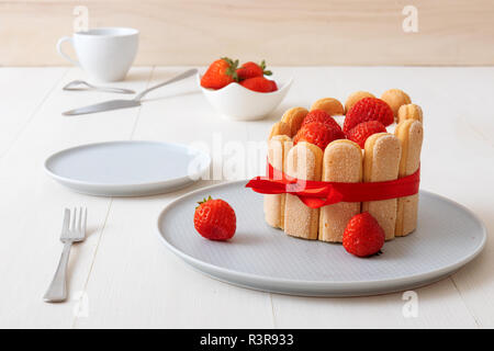 Charlotte, francese dessert con fragole, legato con un nastro rosso scarlatto, tazza di caffè, la ciotola con le fragole sul tavolo bianco. Foto Stock