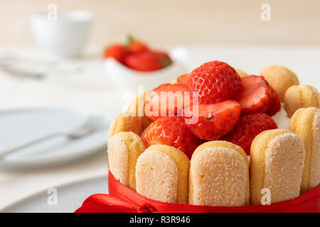 Charlotte, francese dessert con fragole, legato con un nastro rosso scarlatto, tazza di caffè, la ciotola con le fragole sul tavolo bianco. Close up, vista in dettaglio. Foto Stock