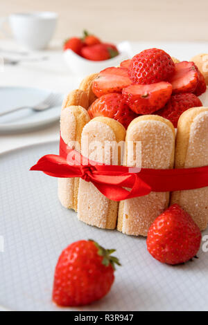 Charlotte, francese dessert con fragole, legato con un nastro rosso scarlatto, tazza di caffè, la ciotola con le fragole sul tavolo bianco. Close up, vista in dettaglio. Foto Stock