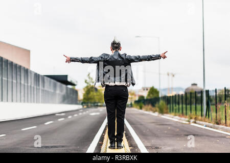 Vista posteriore del punk donna in piedi su una barriera al bordo della strada con le braccia tese Foto Stock