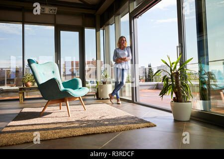 Donna matura in piedi presso la finestra a casa guardando fuori Foto Stock