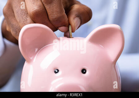 Mano d'uomo inserimento di moneta in Piggybank sulla scrivania in legno Foto Stock