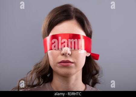 Giovane donna con gli occhi coperti con nastro rosso su sfondo grigio Foto Stock