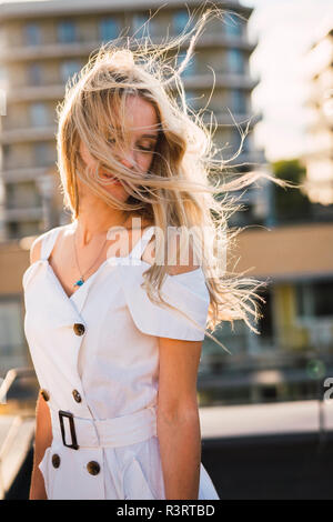 Giovane donna bionda con capelli spazzate dal vento che indossa abito bianco all'aperto Foto Stock