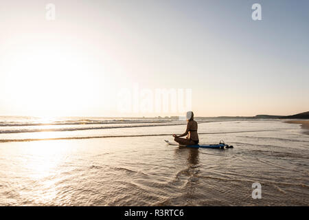 Giovane donna a praticare yoga sulla spiaggia, seduti su una tavola da surf, meditando Foto Stock