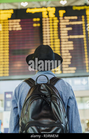 Giovane uomo con il cappello e zainetto guardando arrivo partenza pensione all'aeroporto Foto Stock