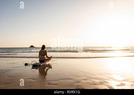 Giovane donna a praticare yoga sulla spiaggia, seduti su una tavola da surf, meditando Foto Stock