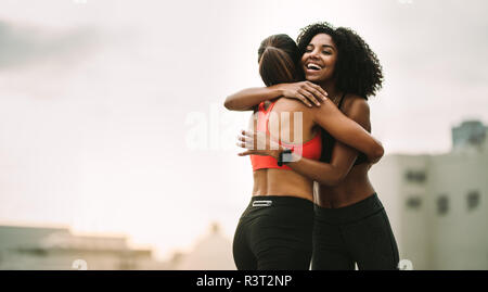 Sorridente afro american donna atleta dando un abbraccio a un altro centro fitness donna in piedi sul tetto. Allegro atlete che abbraccia ogni altro dopo wor Foto Stock