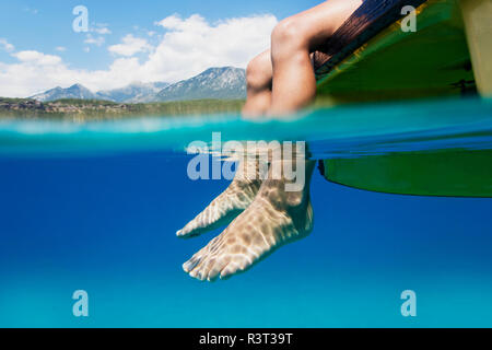 I piedi di un ragazzo seduto sulla barca appesa in acqua Foto Stock