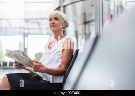 Senior donna seduta in area di attesa con quotidiano Foto Stock