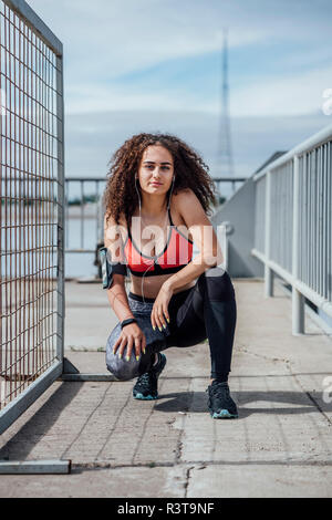 Ritratto di giovane donna atletica con gli auricolari accovacciato all'aperto Foto Stock