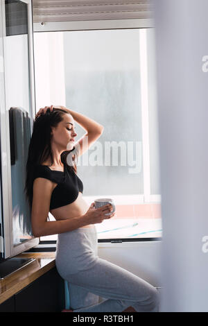 Bella giovane donna con tazza di caffè presso la finestra a casa Foto Stock