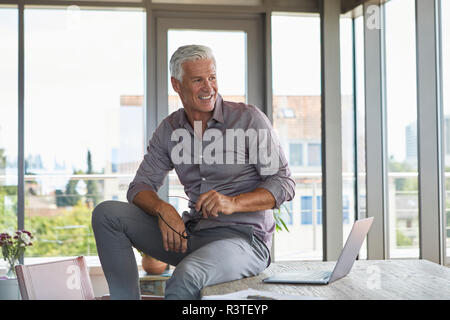 Sorridente uomo maturo con laptop seduto sul tavolo a casa Foto Stock